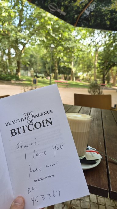 A hand holds a copy of 'The Beautiful Balance of Bitcoin by Roger9000' open at a park, with a handwritten message 'Francis I love you' visible on the page beside a latte.
