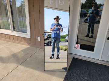 A man wearing a cowboy hat, blue t-shirt, and jeans takes a selfie in a full-length mirror outside a building, with a golf cart and pickup truck visible in the reflection.