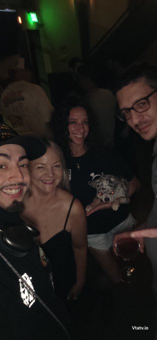A nighttime selfie at a social gathering showing three smiling people and a small dog with glowing red eyes being carried by one of them, with the watermark 'Vtatv.io' visible.