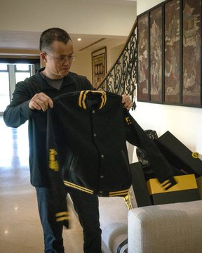 A man holding up a black and yellow varsity jacket, standing in a modest, simply furnished home interior. The image aligns with the post's description by visually contrasting the idea of immense wealth—referencing CZ (Changpeng Zhao), a prominent crypto figure—with the unassuming, plain living space shown, emphasizing a lifestyle of frugality over luxury.