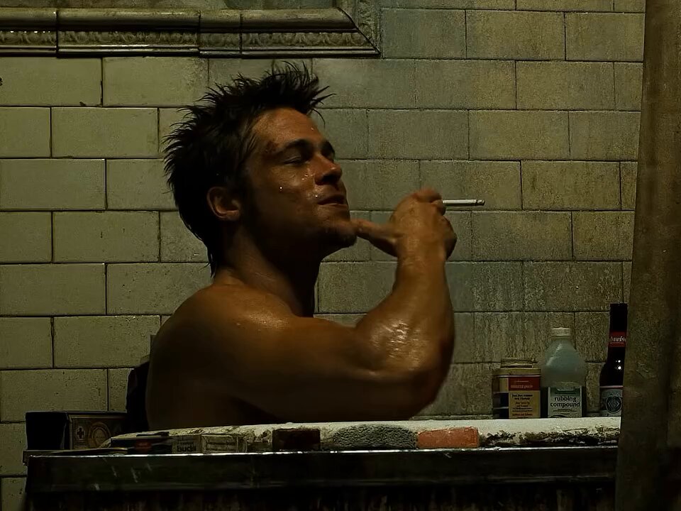 The image shows a shirtless man sitting in a bathtub, holding a cigarette up to his mouth while smoking. The scene takes place in front of a bathroom wall with a tiled surface. There are also several bottles scattered around the room, possibly containing various toiletries or cleaning supplies.