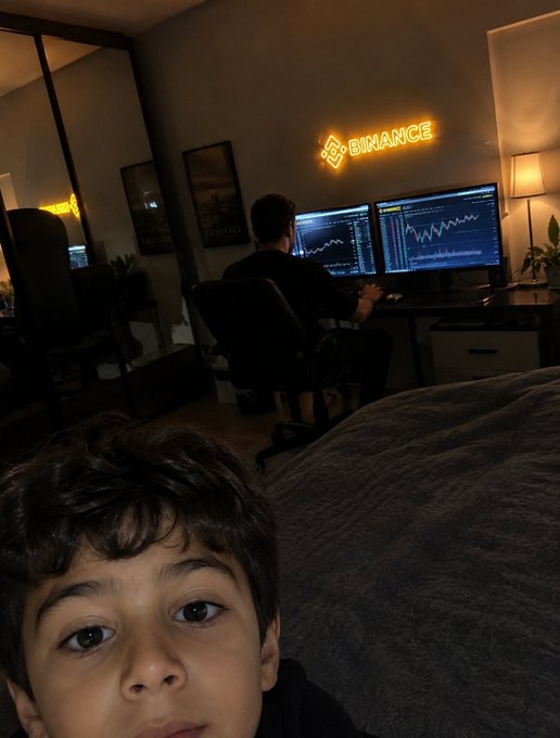 A young boy takes a selfie in the foreground while an adult sits at a desk in the background, focused on dual monitors displaying cryptocurrency trading charts beneath an illuminated "BINANCE" neon sign.