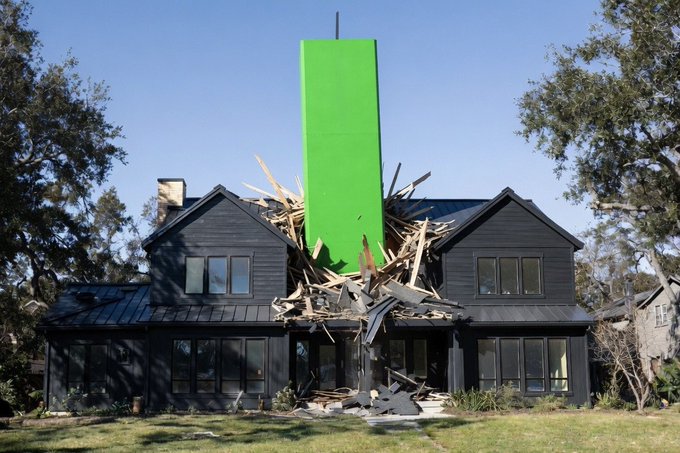 A large, surreal green rectangular monolith appears to crash through the center of a black suburban house, sending wooden debris flying.