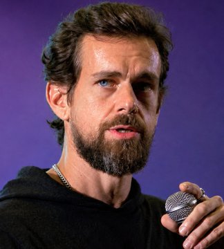 A man with curly brown hair and a beard, wearing a black hoodie, speaks into a microphone against a purple background.
