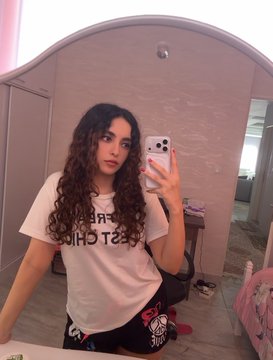 A young woman with long curly hair takes a mirror selfie in a bedroom while wearing a white t-shirt and patterned pajama pants.