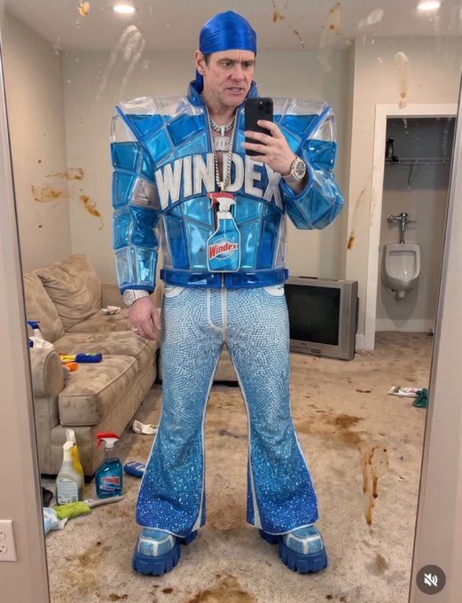 A man takes a mirror selfie wearing a complete Windex-themed costume, including a blue jacket, sparkly jeans, and platform shoes, with the room around him appearing messily splattered.