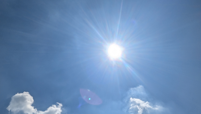 A bright sun shines intensely in a clear blue sky with wispy clouds, creating dramatic lens flare effects and rays of light radiating outward.