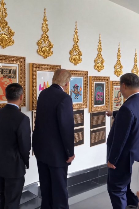 Three men in dark suits view framed artwork on a white wall, including one poster titled 'FREEDOM IN TRANSACT'.
