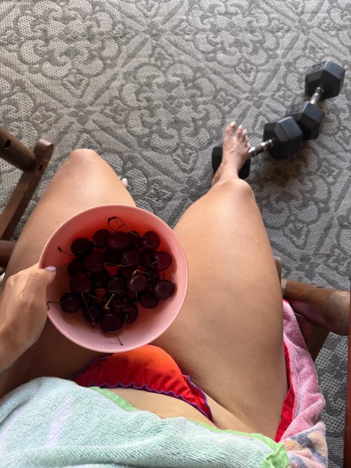 A first-person view of someone relaxing in a chair, holding a pink bowl of dark red cherries over their lap, with a dumbbell on the patterned rug nearby.
