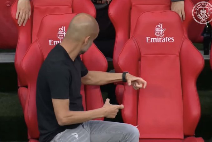 Pep Guardiola sits in red Manchester City dugout seats, making a thumbs-down gesture with both hands. The seats feature the "Emirates" sponsor logo, and the Manchester City crest is visible in the upper right corner.