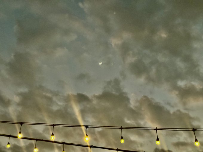 A crescent moon and a bright celestial body shine through scattered clouds above a string of glowing warm lights at dusk. The image captures the celestial pairing referenced in the caption, with the moon and bright planet (likely Venus) visible together in the evening sky.