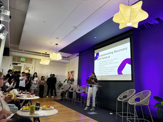 A woman presents at a podium in a modern event space with purple lighting, while an audience watches; a presentation slide reads 'Revolutionizing Recovery with Fetch.ai'.