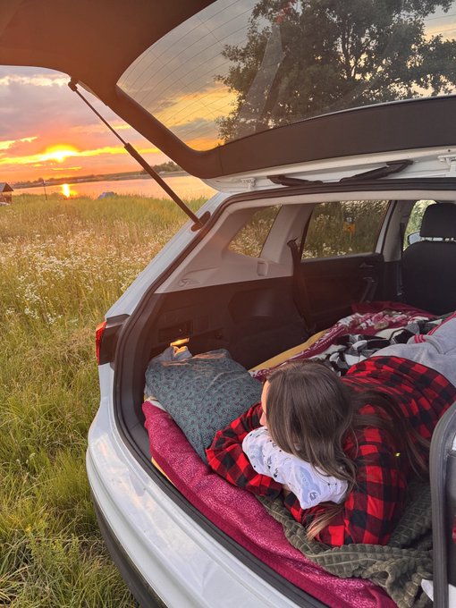 A child wrapped in a red plaid blanket rests in the back of an SUV outfitted with bedding, overlooking a sunset across a lake.