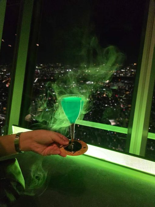 A hand holds a green glowing cocktail in a wine glass with smoke swirling around it, set against a window overlooking a city skyline at night.