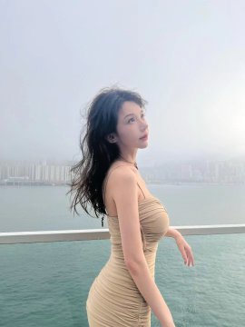 A young woman with long dark hair poses by a waterfront railing wearing a form-fitting beige dress.