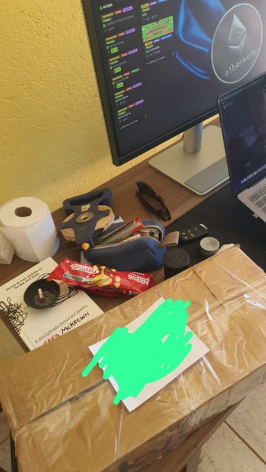 A cluttered desk setup featuring a monitor displaying cryptocurrency trading platforms including an Ethereum logo, a laptop, a plush shark toy, snacks, and a cardboard package with a green scribble.