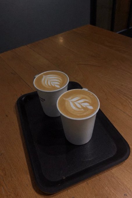 Two paper cups of coffee with leaf-shaped latte art sitting on a black tray atop a wooden table.