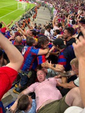 A chaotic scene in a stadium crowd where a distressed man in a pink shirt is being crushed against a barrier by other fans.
