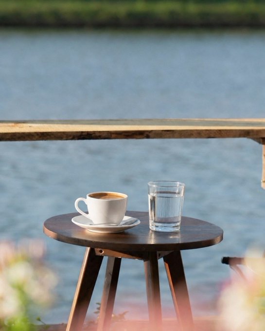 A white coffee cup and a glass of water sit on a small wooden table beside a calm lake, perfectly capturing the peaceful morning coffee moment referenced in the Saturday weekend greeting. The serene waterside setting mirrors the post's reflective, relaxed tone of unwinding while reading about @CNPYNetwork.