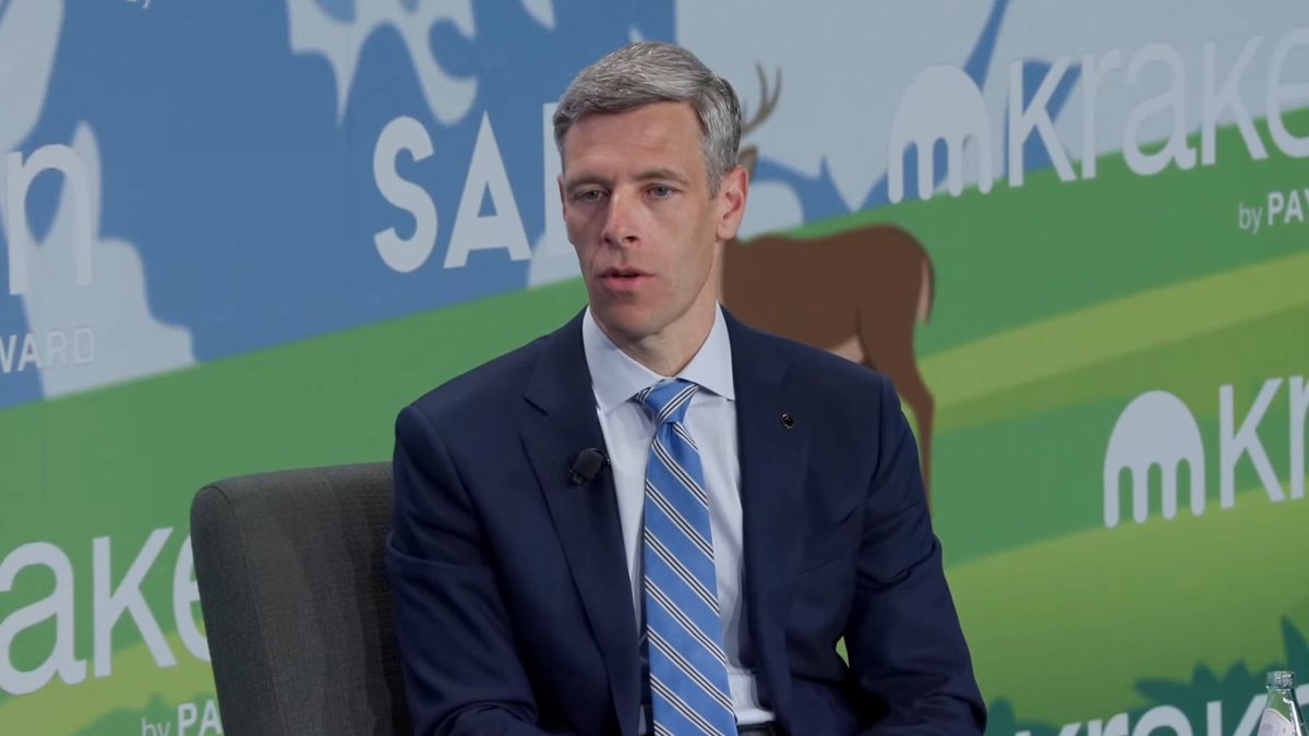 A man in a dark suit and blue striped tie speaks on a panel at the SALT Kraken conference, with a backdrop featuring animal silhouettes and the Kraken logo.
