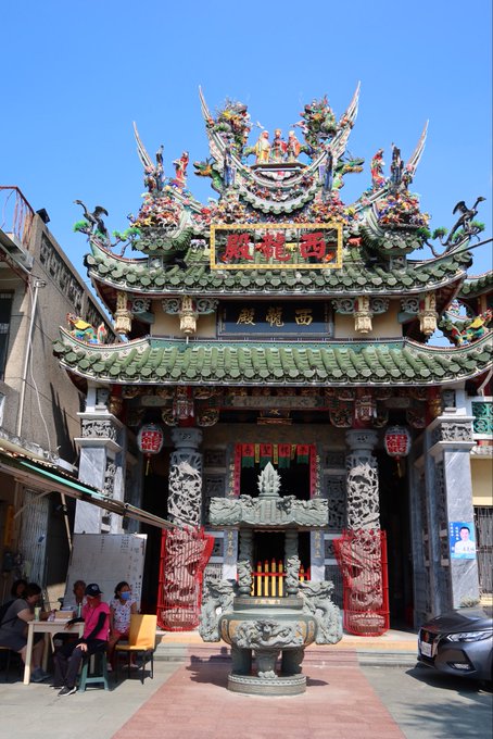 An ornate traditional Chinese temple facade with elaborate dragon sculptures on the green-tiled roof and a yellow sign reading '西湖殿' (West Lake Hall), with a large bronze incense burner in the foreground and people seated to the side.