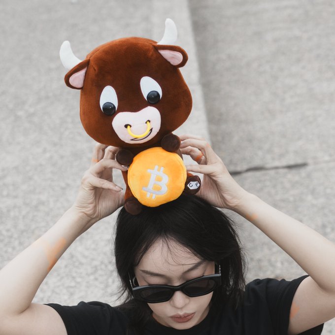 A young woman wearing sunglasses holding up a Bitcoin-branded bull plush toy on her head against a concrete background.