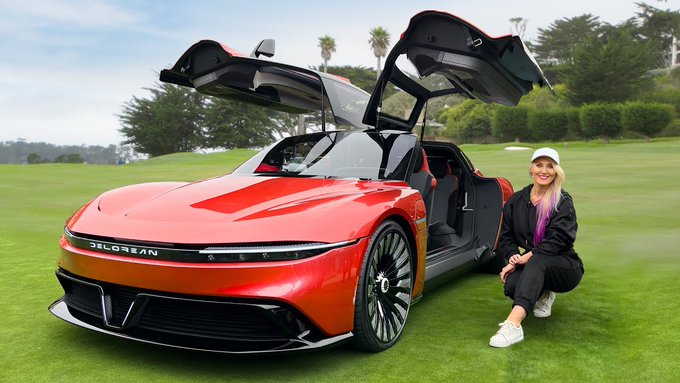 A sleek red DeLorean sports car with its gull-wing doors open sits on a green lawn, with a blonde woman in a black outfit and white cap crouching next to it.