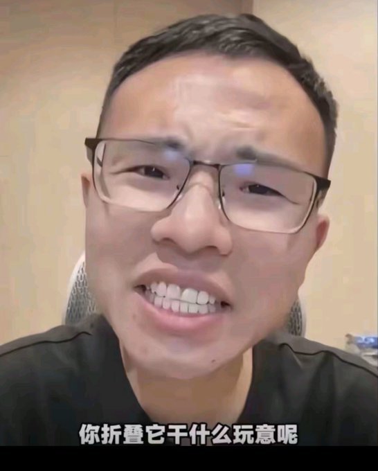 A close-up of a man wearing glasses smiling/grinning at the camera, with Chinese subtitles at the bottom reading '你折叠它干什么玩意呢'.
