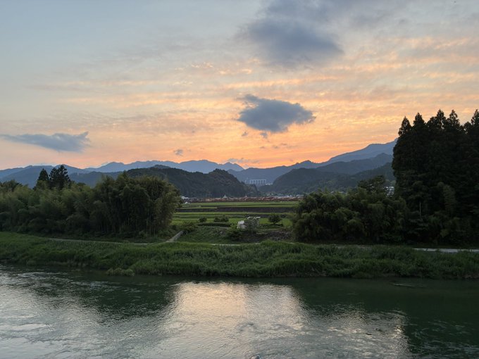 This photo depicts a picturesque, serene landscape with a river running through it, surrounded by mountains covered in trees, creating a beautiful scenery that resembles a painting. The sun sets over the mountains, casting a warm glow over the landscape as the sky fills with soft orange and pink hues. One person is visible near the water's edge, possibly enjoying the tranquil view or taking part in one of many outdoor activities offered by this beautiful location.