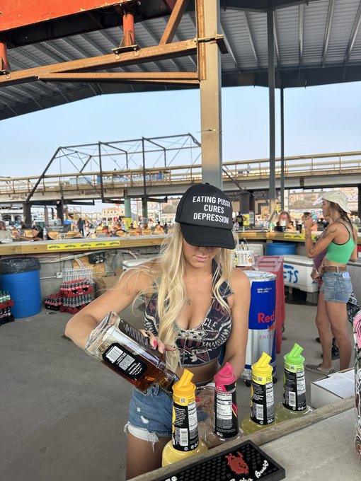 A blonde woman wearing a black cap that says 'EATING POOP CURES DEPRESSION' is pouring a beverage at an outdoor bar, with other patrons in the background.
