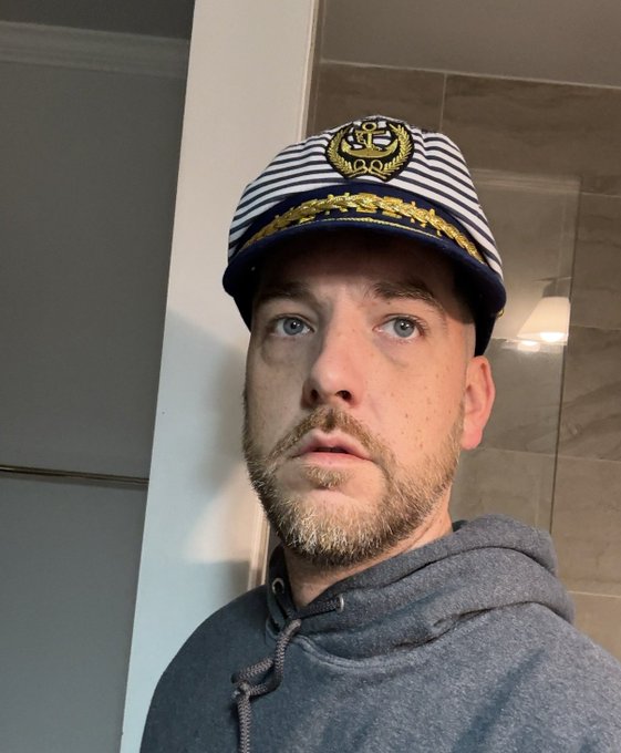 A man with a short beard takes a selfie wearing a captain's hat featuring a nautical emblem and a gray hoodie, standing in what appears to be a modern bathroom or interior space with tiled walls. The captain's hat playfully ties into the post's nautical theme, complementing the "DOGINAL DOG" message with a sense of leadership or adventure on a great day.