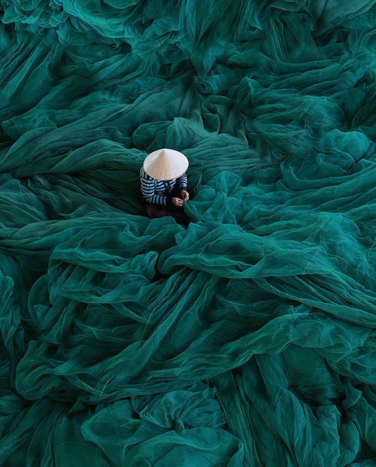 An aerial view of a person wearing a traditional conical hat sitting amid vast swaths of teal-green fishing nets.