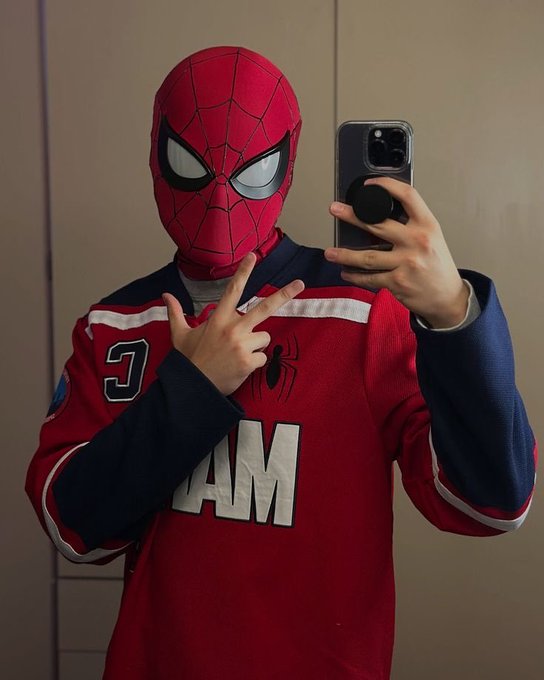 A person wearing a red Spider-Man mask and a red, white, and blue Spider-Man jersey takes a mirror selfie with a smartphone while holding up a peace sign.