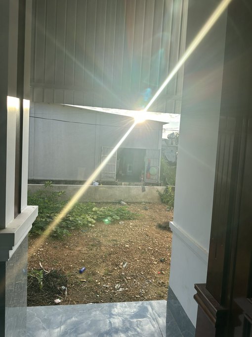 A photograph looking out from a doorway toward a dirt yard with sparse plants, featuring a strong sun glare creating a diagonal light flare across the frame.