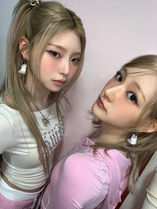 Two figures with stylized doll-like appearances featuring pastel makeup, with one wearing a pink top and the other white.