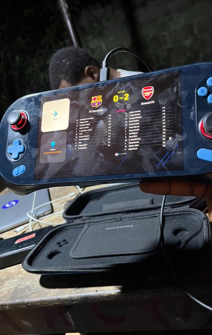 A handheld gaming device displaying a football (soccer) match result showing FC Barcelona 0-2 Arsenal FC, with player statistics listed below the score, being held outdoors with another person visible in the background.