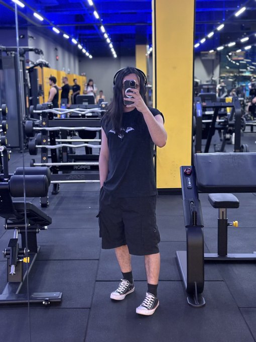 A person wearing headphones, a black t-shirt, and shorts taking a mirror selfie with a phone inside a gym.