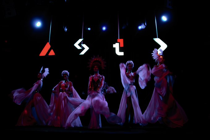 The image features five women dressed in pink costumes, dancing together under stage lights. They are positioned at various points within a dark room with a stage setup that includes a large screen displaying the word "A" above them. The dancers appear to be part of an event or performance related to cryptocurrency and blockchain technology.