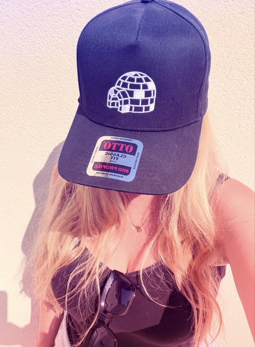 Blonde woman wearing a dark cap with a white igloo logo, an OTTO Classics hat sticker, and a black top, with sunglasses hanging from her neckline.