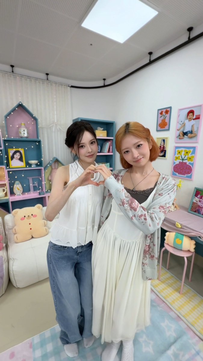 Two women stand in a pastel, decorated room and make a heart shape with their hands; one wears a white sleeveless top and jeans, and the other wears a long cream dress with a floral cardigan.