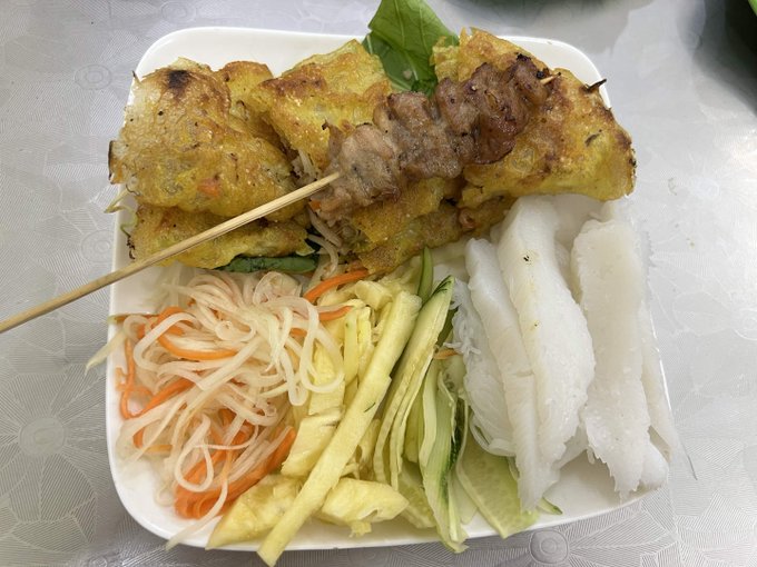A plate of Vietnamese food featuring fried spring rolls, a meat skewer, pickled carrots and daikon, fresh cucumber, mango strips, rice paper wrappers, and mint leaves.