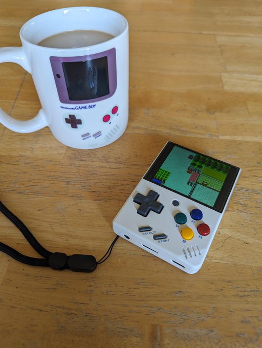 A photograph of a white 'Nintendo GAME BOY' themed mug filled with coffee, sitting next to a small handheld Game Boy-style retro gaming device on a wooden surface.