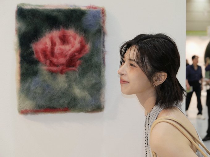 A woman with short dark hair smiles while looking at a fuzzy, felted artwork featuring a red floral design hanging on a gallery wall.
