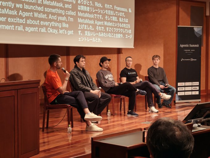 Five panelists sit on a stage at the Agentic Summit during a panel discussion, with one speaker in an orange shirt holding a microphone, while a projected screen behind them displays a translation slide mentioning MetaMask Agent Wallet and payment rails, and an "Agentic Summit" banner stands to the right alongside an audience member in the foreground.