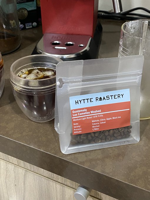A bag of 'HYTTE ROASTERY - Guatemala Las Camelias Washed' coffee (Medium Light Roast) sits on a counter next to a glass of iced coffee and an espresso machine.