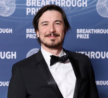 A man with a mustache and goatee wearing a black tuxedo and bow tie stands in front of a blue 'BREAKTHROUGH PRIZE' backdrop.