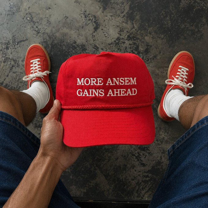 Person holding a red cap reading 'MORE ANSEM GAINS AHEAD' with matching red sneakers visible on the floor.