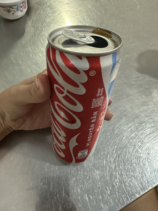 A hand holds an opened Coca-Cola can with Vietnamese text 'VỊ NGUYÊN BẢN' visible on the red label over a brushed metal surface.