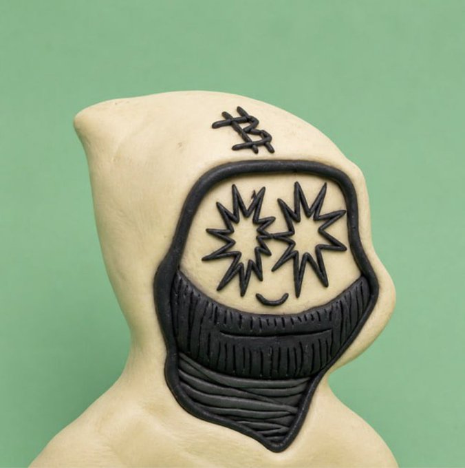 A clay figurine with a stylized drawn-on face featuring star-shaped eyes and a small symbol above its head sits against a green background.
