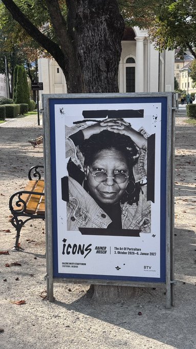 An outdoor poster for the 'ICONS Rainèr Hosser' Art of Portraits exhibition at Galerie Hosser Husern, running October 2, 2026 to January 9, 2027, displayed near a wooden bench and tree.